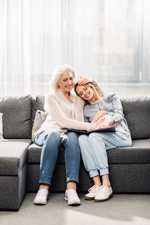 Young pregnant woman looking at photo album with mother on gray sofa in roomの写真素材
