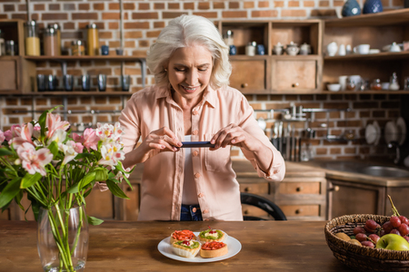 Portrait of senior woman photographing plate with food at kitchenの写真素材