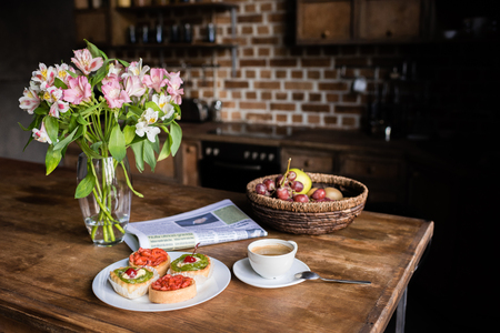 Still life of flowers, newspaper, breakfast with cakes, coffee and fruits on kitchen table 