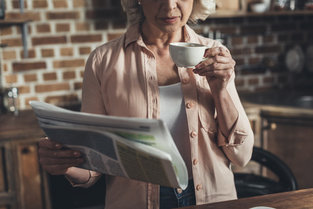 Portrait of senior woman reading newspaper and drinking coffee during breakfast in kitchenの写真素材