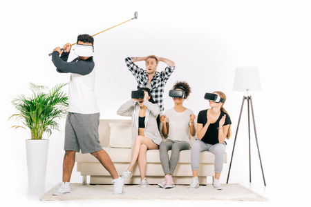 emotional young multiethnic people in virtual reality headsets looking at friend playing golf isolated on whiteの写真素材