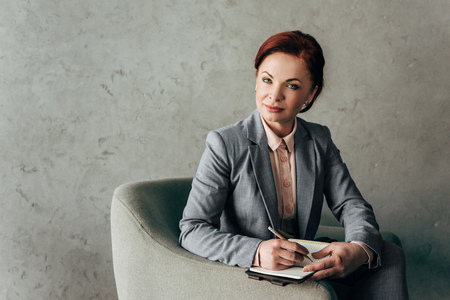confident mature businesswoman with notebook sitting in armchair on concrete wall backgroundの写真素材
