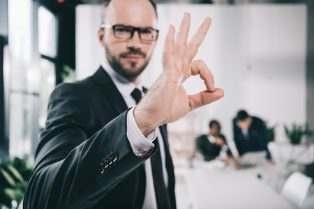 handsome young businessman showing okay sign with blurred colleagues on backgroundの写真素材
