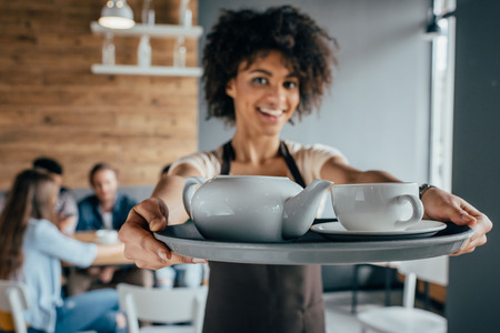 Smiling african american waitress holding tray with tea and customers sitting behind her in cafeの写真素材