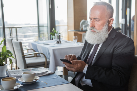 mature bearded businessman using smartphone in restaurant while waiting for meetingの写真素材