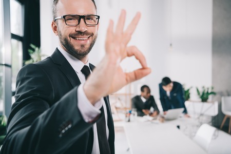 handsome young businessman showing okay sign with blurred colleagues on backgroundの写真素材