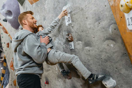 Young dad teaching his little son how to climb a wall with grips at gymの写真素材