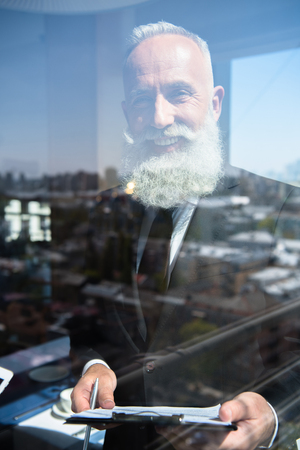 smiling mature businessman with clipboard behind window on sunny dayの写真素材