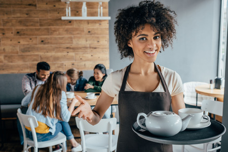 Smiling african american waitress holding tray with tea and customers sitting behind her in cafeの写真素材