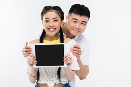 happy young asian couple showing digital tablet with blank screen and smiling at camera isolated on whiteの写真素材