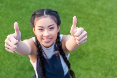 high angle view of smiling asian girl showing thumbs up while standing on green lawn  の写真素材