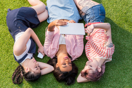 top view of three multiethnic girls using digital tablet while lying on green grass