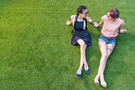 beautiful happy multiethnic girls in sunglasses smiling each other while sitting on grass の写真素材