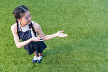 smiling asian girl gesturing with hands and showing copy space above green lawnの写真素材