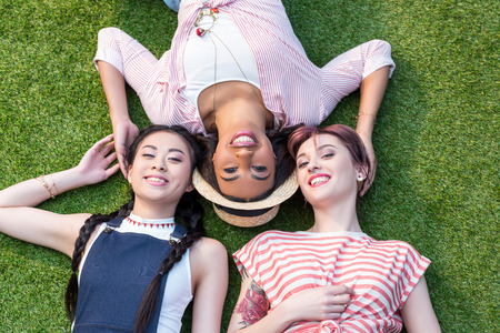 top view of beautiful happy multiethnic girls smiling at camera while lying on lawn