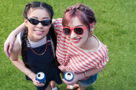 high angle view of young multiethnic women in sunglasses holding soda cans and smiling at camera の写真素材