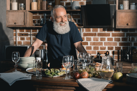 portrait of smiling senior man leaning on table and looking away in kitchen at homeの写真素材