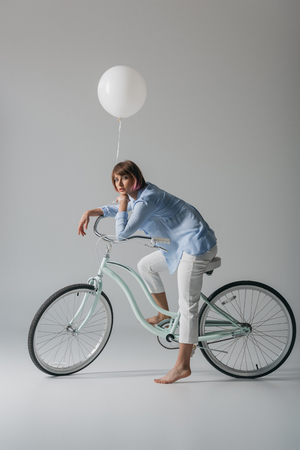 happy trendy girl sitting on bike with white balloon, isolated on greyの写真素材