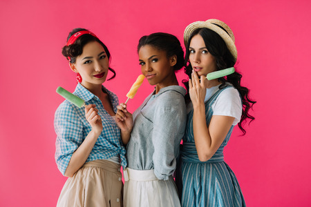 portrait of multicultural stylish women with popsicles looking at camera isolated on pinkの写真素材