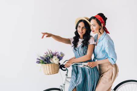 portrait of multicultural women in stylish retro clothing sitting on bicycle isolated on greyの写真素材
