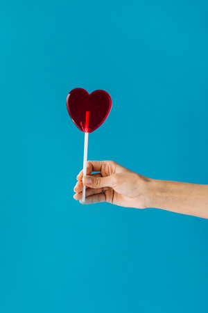 cropped shot of female hand holding heart shaped lollipop isolated on blueの写真素材