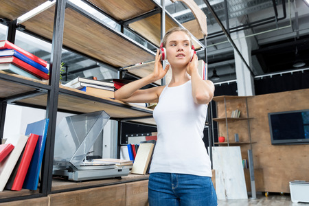 Young attractive woman wearing headphones and listening to music next to bookshelf in living room