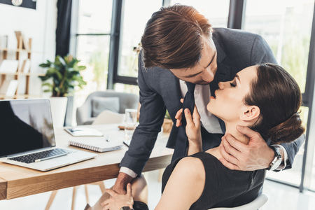 Young man in a suit standing over beautiful woman sitting in a chair and leaning in for a kissの写真素材