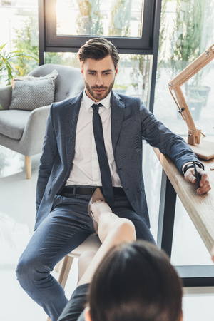 Woman in high heels placing her foot on a chair between legs of young man in business suitの写真素材
