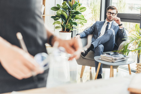 Young businessman in suit sitting in armchair at office and looking at his secreteryの写真素材