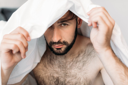 handsome shirtless man hiding under blanket and looking at cameraの写真素材