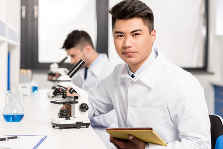Young scientist in lab coat sitting at table in laboratory with microscope and digital tablet and...