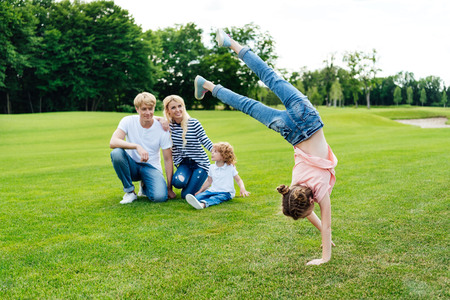 happy parents with son looking at cute little daughter performing handstand at park