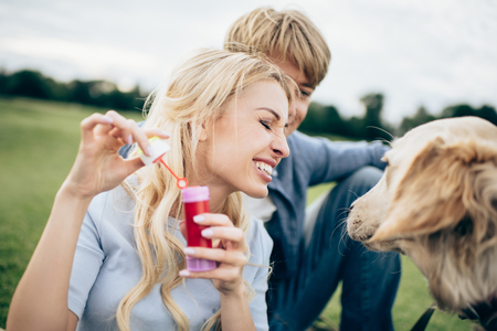 happy young couple blowing soap bubbles while resting with dog at parkの写真素材