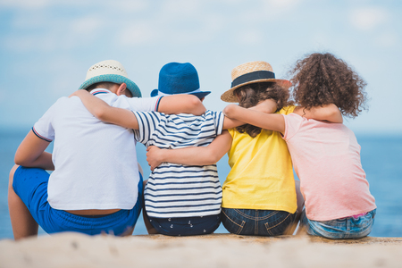 back view of boys and girls sitting together on wooden trunk at seasideの写真素材