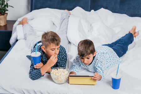 little boys eating popcorn and drinking soda while resting in bedの写真素材
