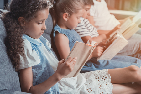 multicultural group of focused children reading books while sitting on sofa at home togetherの写真素材