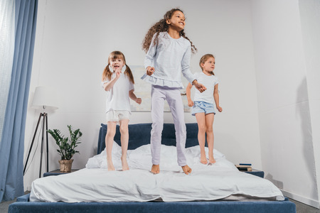 multiethnic little girls in pajamas jumping on bed at homeの写真素材