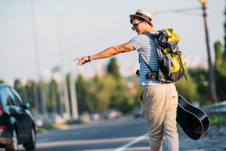 back view of young man with guitar gesturing to stop car while hitchhiking aloneの写真素材