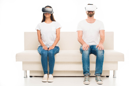 'Portrait of young happy couple sitting on sofa using virtual reality headsets, isolated on white