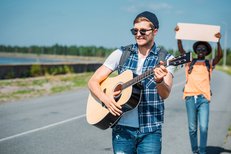 selective focus of caucasian man playing guitar while african american friend with empty cardboard hitchhiking behindの写真素材