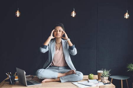 african american businesswoman sitting in lotus pose on table while meditating in office の写真素材