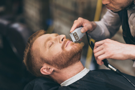 Male barber trimming customers beard in barber shop の写真素材