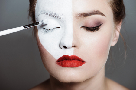 young beautiful teen girl applying creative white bodyart with makeup brush on face, isolated on grey の写真素材