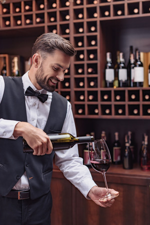 Cropped view sommelier pouring red wine from bottle into glass at table in cellarの写真素材