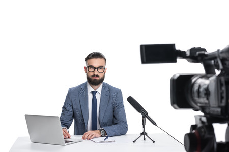 male newscaster sitting at table with laptop, notepad and microphone and looking at camera, isolated...