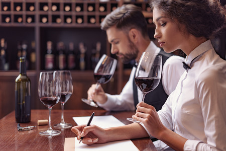 Two sommeliers, male and female tasting red wine and making notes at degustation card