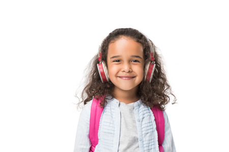 adorable african american schoolgirl listening music in headphones and smiling at camera isolated on white の写真素材
