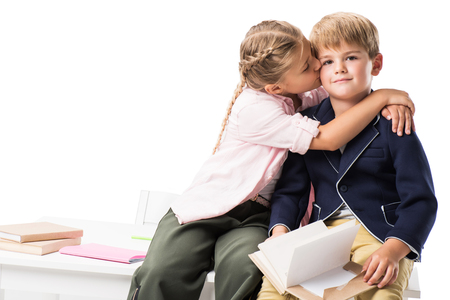 cute little girl kissing boy holding book and looking at camera isolated on whiteの写真素材