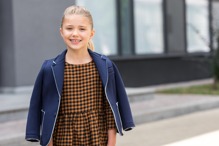 portrait of adorable little blonde girl smiling at camera while standing near schoolの写真素材