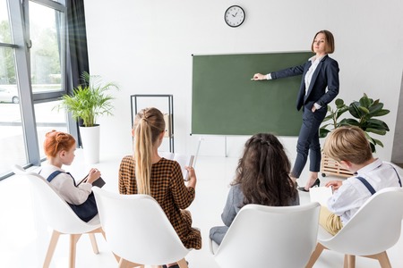 young female teacher holding chalk and pointing at blackboard while explaining lesson to students の写真素材